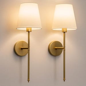 May include: Two wall sconces with a gold finish. Each sconce has a circular base, a long, slender stem, and a white lampshade. The lampshades are conical and emit a warm, inviting glow. The sconces are mounted on a light-colored wall.