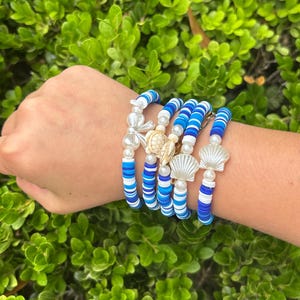 May include: A collection of blue and white beaded bracelets with seashell and pearl accents. The bracelets feature various shades of blue and white beads, with decorative seashell and pearl charms. The bracelets are displayed on a wrist.