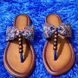 Könnte beinhalten: Ein Paar braune Ledersandalen mit blauem Zehenriemen und einer dekorativen Schleife. Die Schleife besteht aus gemustertem Stoff mit roten, blauen und orangefarbenen Paisley-Designs. Holzperlen schmücken die Mitte der Sandalen.