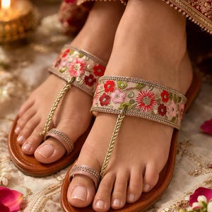 May include: Close-up of brown leather sandals with floral embroidery. The sandals feature a gold chain toe loop and a strap adorned with red, pink, and green flowers. The sandals are on a decorative surface.