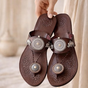 May include: A pair of brown leather sandals with embossed floral patterns. The sandals have silver embellishments, including circular medallions and small elephant figures. The thong straps are decorated with silver elements. The sandals are made of leather.