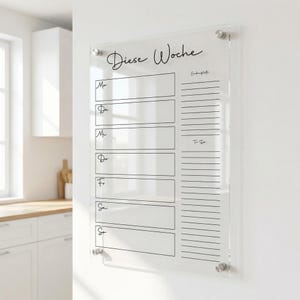 May include: A clear acrylic weekly planner, with the words "Diese Woche" in script. The planner has sections for each day of the week, plus a to-do list. It is mounted on a white wall with silver fixings.