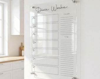 May include: A clear acrylic weekly planner with the words "Diese Woche" in script. The planner has sections for each day of the week, plus a to-do list. It is mounted on a white wall with silver hardware.
