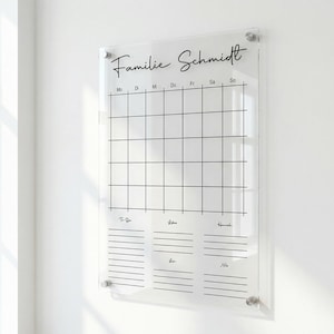 Könnte beinhalten: Ein klarer Wandkalender aus Acryl mit dem Text "Familie Schmidt" in schwarzer Schrift. Das Kalendarium ist in Wochentage unterteilt und bietet Platz für Notizen. Er ist an einer weißen Wand mit silberner Hardware befestigt.