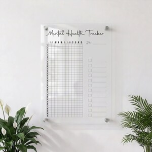 Könnte beinhalten: Ein klarer Mental Health Tracker aus Acryl mit schwarzer Schrift. Der Tracker hat ein Raster für tägliche Einträge und Kontrollkästchen. Die Monate des Jahres sind oben aufgeführt. An einer weißen Wand montiert.