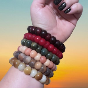 May include: A collection of seven beaded bracelets in various colors, including shades of brown, red, green, and beige. The bracelets are stacked on a wrist, showcasing the different hues and textures of the beads. The background has a gradient of blue, orange, and yellow.