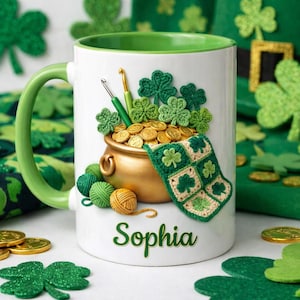 May include: White ceramic mug with a green handle and rim, decorated with a gold pot of coins, crochet shamrocks, and yarn balls. The mug has the name "Sophia" in green script. A St. Patrick's Day theme.