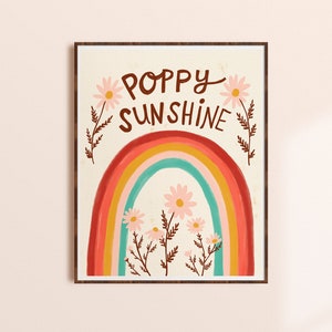 May include: A framed art print featuring a colorful rainbow with the words "Poppy Sunshine" in brown script. The artwork includes daisies and floral accents, set against a cream background. The print is displayed on a light beige wall.