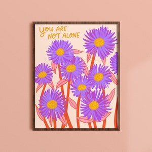 May include: A framed print with a pink background and a cluster of purple flowers with orange centers. The text "You are not alone" is written in orange at the top of the print.