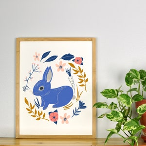 May include: A framed print of a blue rabbit surrounded by a wreath of flowers and leaves. The rabbit is sitting on its hind legs and has a carrot behind it. The flowers are pink, white, and orange.