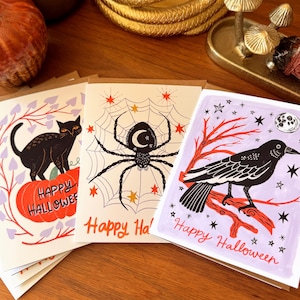 May include: Three rectangular greeting cards featuring Halloween illustrations. One shows a black cat on a pumpkin, one a spider on a web, and one a crow on a branch. Each card includes the text Happy Halloween in a script font with stars.