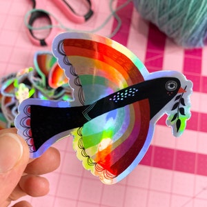 May include: A holographic sticker of a black bird with a rainbow wingspan and an olive branch in its beak. The bird is flying against a pink background.