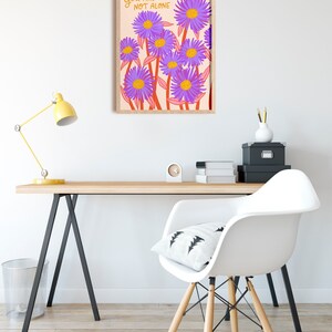 May include: A framed print with a light pink background and purple flowers with yellow centers. The text "YOU ARE NOT ALONE" is written in orange on the print.