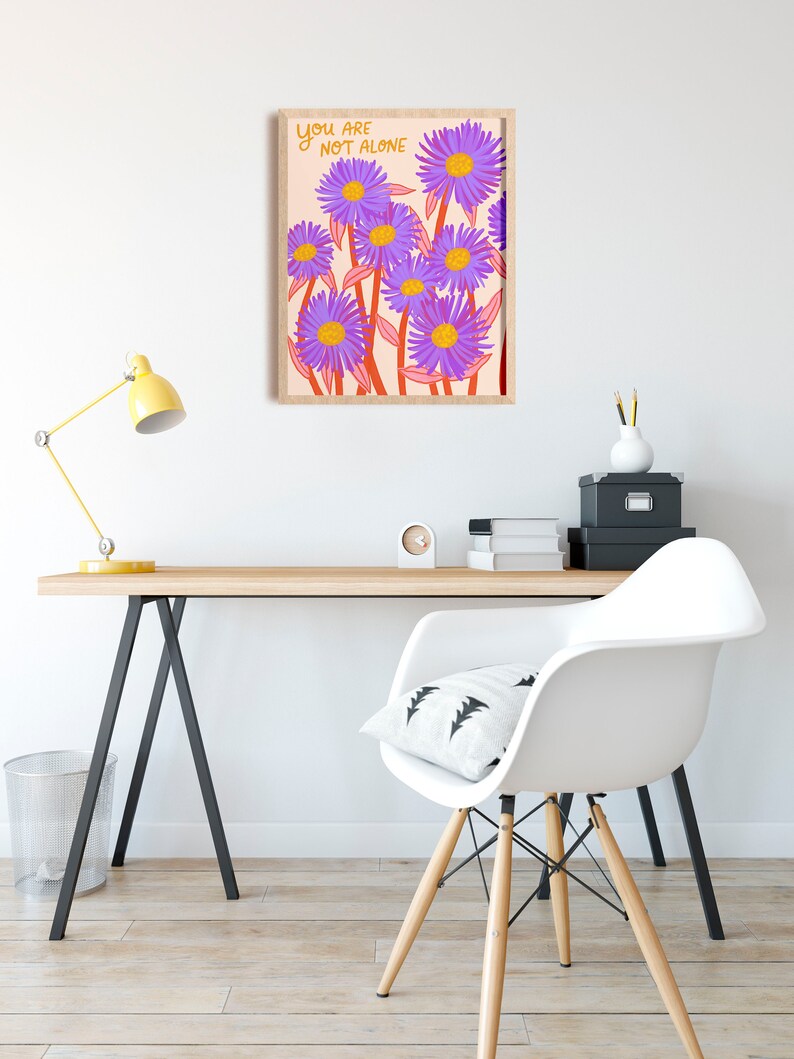 May include: A framed print with a light pink background and purple flowers with yellow centers. The text "YOU ARE NOT ALONE" is written in orange on the print.