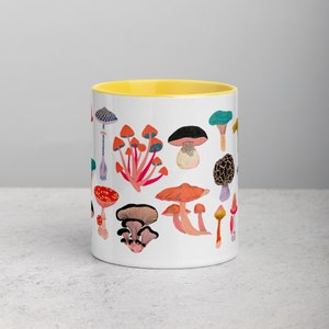 May include: White ceramic mug with a yellow rim. The mug features a colourful illustration of various mushrooms with red, orange, yellow, green, blue, and purple accents.