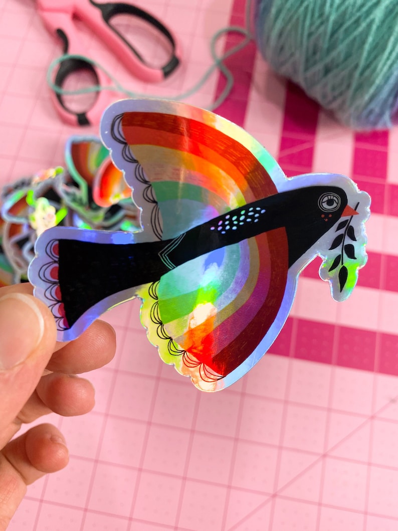 May include: A holographic sticker of a black bird with a rainbow wingspan and an olive branch in its beak. The bird is flying against a pink background.