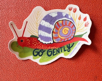 May include: A die-cut sticker featuring a red snail with a purple and orange spiral shell. The snail rests on a green leaf with stylized red and yellow plants in the background. The text GO GENTLY is printed in dark blue script below the snail.