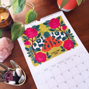 May include: A calendar page for June featuring a colorful illustration of a Garden Tiger Moth surrounded by pink roses. The moth has orange wings with black spots and a white and black pattern. The calendar is on a wooden surface.