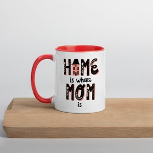 K&ouml;nnte beinhalten: Wei&szlig;e Keramiktasse mit rotem Rand und Henkel. Die Tasse hat eine schwarz-wei&szlig;e Grafik mit dem Text "Home is where Mom is" und einer stilisierten Haus-Illustration.