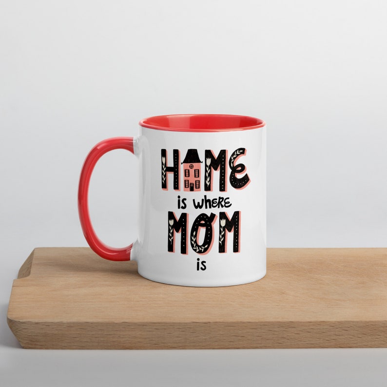 K&ouml;nnte beinhalten: Wei&szlig;e Keramiktasse mit rotem Rand und Henkel. Die Tasse hat eine schwarz-wei&szlig;e Grafik mit dem Text "Home is where Mom is" und einer stilisierten Haus-Illustration.