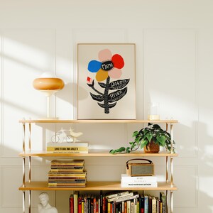 May include: A framed art print featuring a colorful flower design with the words "Mama," "Dakota," and "River." The print is displayed on a wooden shelf with a lamp, books, and decorative items. The color palette includes pink, red, blue, and yellow.