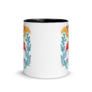May include: White ceramic mug with a black interior. The mug features a colorful design of red mushrooms, yellow flowers, and blue foliage. The words "I SEE MAGIC" are printed on the side of the mug.