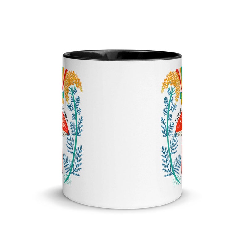 May include: White ceramic mug with a black interior. The mug features a colorful design of red mushrooms, yellow flowers, and blue foliage. The words "I SEE MAGIC" are printed on the side of the mug.