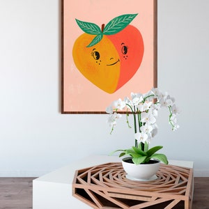 May include: A framed print of a peach with a smiling face. The peach is split in half, with one side yellow and the other side red. The peach is on a pink background. A white orchid plant is in a white pot on a wooden table with a geometric design.