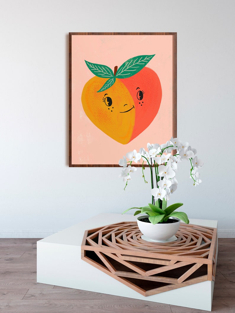May include: A framed print of a peach with a smiling face. The peach is split in half, with one side yellow and the other side red. The peach is on a pink background. A white orchid plant is in a white pot on a wooden table with a geometric design.