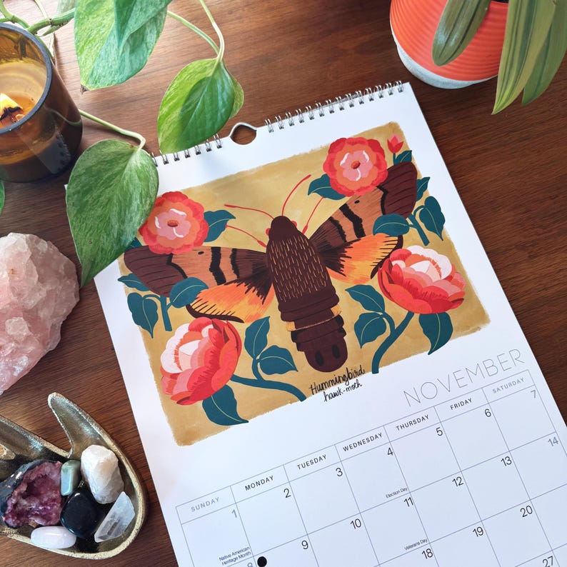 May include: A spiral-bound calendar open to November, featuring a colorful illustration of a hummingbird hawk-moth surrounded by red roses and green leaves. The calendar is on a wooden surface, with other decorative items.