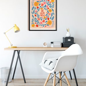May include: A framed print with a floral design and the text "BE HERE NOW" in blue and red lettering on a peach background.