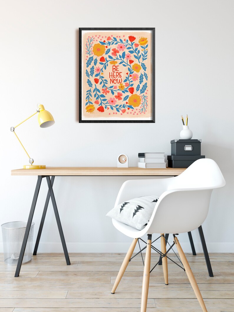 May include: A framed print with a floral design and the text "BE HERE NOW" in blue and red lettering on a peach background.