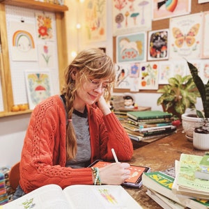 K&ouml;nnte beinhalten: Eine Frau in einem roten Cardigan und einer Brille zeichnet auf einem Tablet. Sie ist umgeben von B&uuml;chern und Kunstwerken mit Natur- und Pflanzenmotiven. Die B&uuml;cher handeln von essbaren Pflanzen und Wildpflanzen.