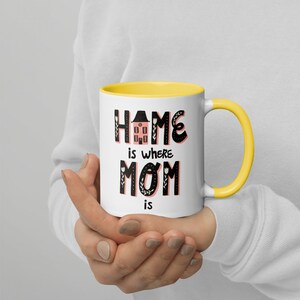 K&ouml;nnte beinhalten: Eine wei&szlig;e Keramiktasse mit gelbem Rand. Die Tasse hat ein schwarzes und pinkfarbenes Design mit dem Text "Home is where Mom is".