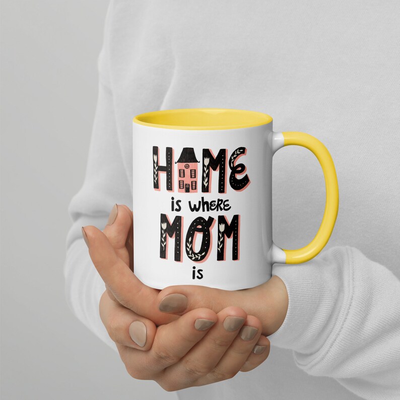 K&ouml;nnte beinhalten: Eine wei&szlig;e Keramiktasse mit gelbem Rand. Die Tasse hat ein schwarzes und pinkfarbenes Design mit dem Text "Home is where Mom is".