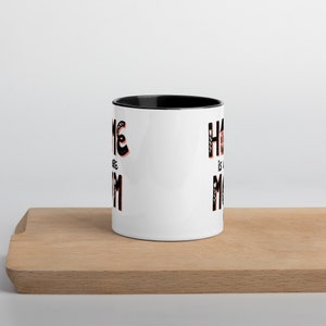K&ouml;nnte beinhalten: Wei&szlig;e Keramiktasse mit schwarzem Rand. Die Tasse hat ein schwarz-wei&szlig;es Motiv mit dem Text "Home is where the Mom is".