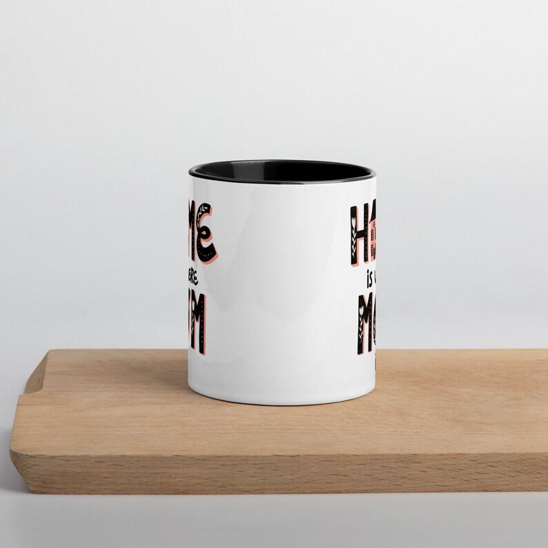 K&ouml;nnte beinhalten: Wei&szlig;e Keramiktasse mit schwarzem Rand. Die Tasse hat ein schwarz-wei&szlig;es Motiv mit dem Text "Home is where the Mom is".