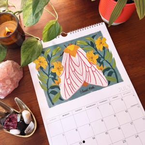 May include: A spiral-bound calendar open to July, featuring a colorful illustration of a Ghost Moth with white wings and red veins, surrounded by yellow flowers. The calendar is on a wooden surface, with a lit candle and plants in the background.