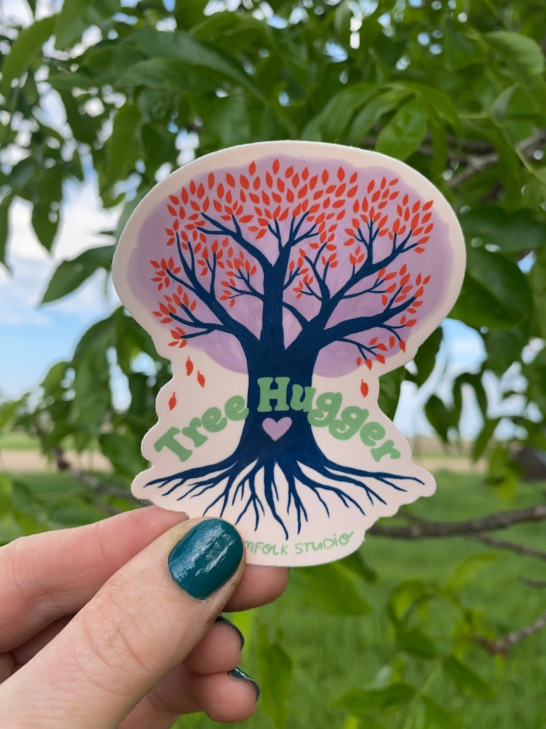 May include: A sticker featuring a stylized tree with red leaves and blue roots. The text "Tree Hugger" is written in green letters around the tree. The sticker is held in a hand with green nail polish.