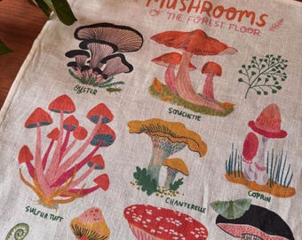 May include: A beige linen tea towel featuring a colorful botanical illustration titled MUSHROOMS OF THE FOREST FLOOR. It displays various mushroom species including Oyster, Souchette, Sulfur Tuft, Chanterelle, Coprin, and Porcini with detailed labels.