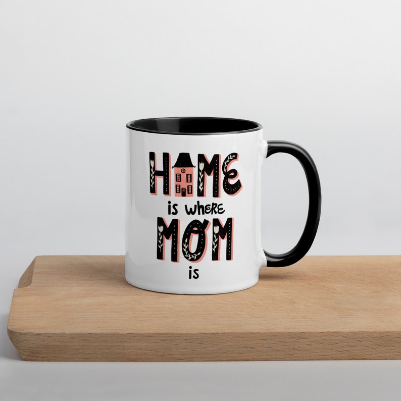K&ouml;nnte beinhalten: Wei&szlig;e Keramiktasse mit schwarzem Rand und Henkel. Die Tasse hat eine schwarze Grafik mit der Aufschrift "Home is where Mom is" mit einer kleinen Haus-Illustration in der Mitte des Wortes "Home".
