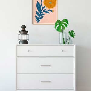 K&ouml;nnte beinhalten: Eine wei&szlig;e Kommode mit drei Schubladen und einem gerahmten Druck einer stilisierten orangefarbenen Blume mit blauen Bl&auml;ttern auf rosa Hintergrund. Die Kommode steht auf einem Holzboden.