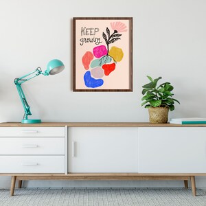 May include: A framed print with a pink background and a colorful abstract illustration of a plant with the text "KEEP growing". The print is hanging on a white wall above a white dresser with three drawers and two doors.