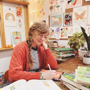 May include: A woman wearing a red cardigan and glasses is drawing on a tablet. She is surrounded by books and artwork featuring nature and plants. The books are about edible plants and wild plants.