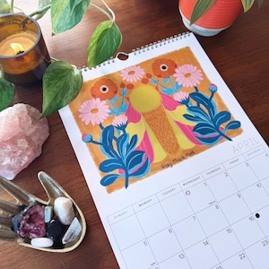 May include: A spiral-bound calendar open to April, featuring a colorful floral illustration with orange, pink, and blue hues. The calendar is on a wooden surface, with a candle, plants, and crystals nearby. The artist's signature is visible.