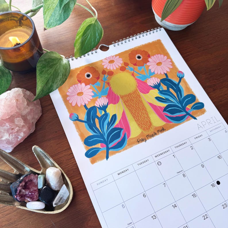 May include: A spiral-bound calendar open to April, featuring a colorful floral illustration with orange, pink, and blue hues. The calendar is on a wooden surface, with a candle, plants, and crystals nearby. The artist's signature is visible.