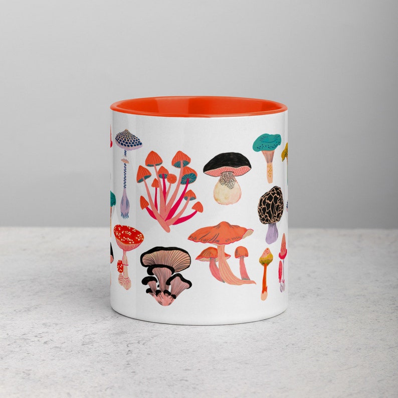 May include: White ceramic mug with an orange rim featuring a colourful illustration of various mushrooms. The mushrooms are depicted in different shapes, sizes, and colours, including red, orange, yellow, green, blue, and purple.