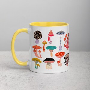 May include: White ceramic mug with a yellow rim featuring a colourful illustration of various mushrooms. The mug is on a white surface.