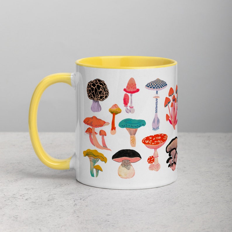 May include: White ceramic mug with a yellow rim featuring a colourful illustration of various mushrooms. The mug is on a white surface.