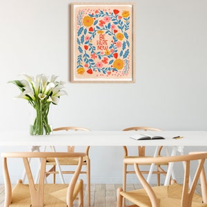 May include: A framed print with a floral design and the text "BE HERE NOW" in red and white lettering. The flowers are in shades of blue, pink, yellow, and orange.